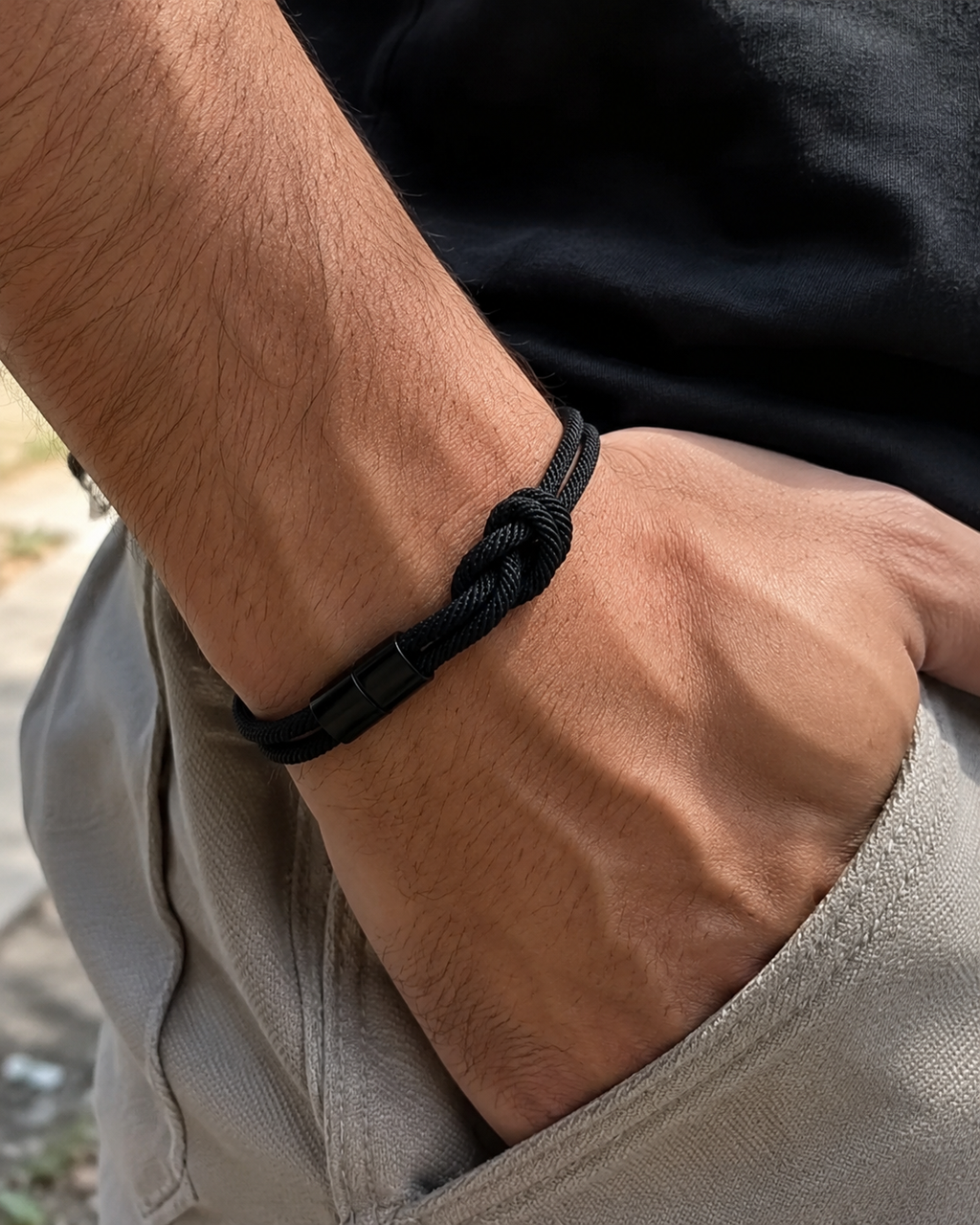A close-up of a person's wrist wearing a black braided cord bracelet with a simple black buckle, paired with a black t-shirt and beige pants.